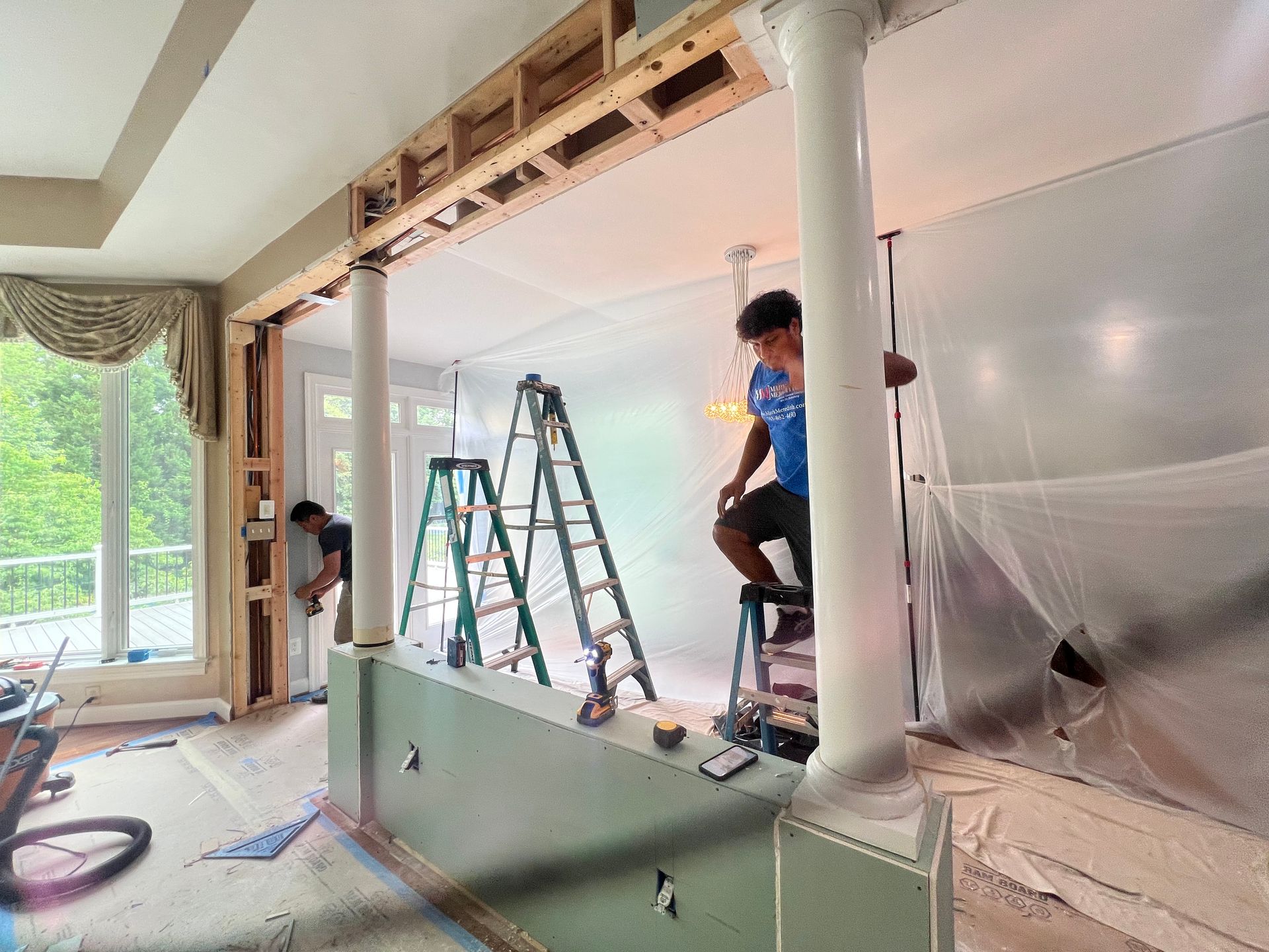 Workers installing a partition wall in a bright room, with ladders, tools, and plastic sheeting