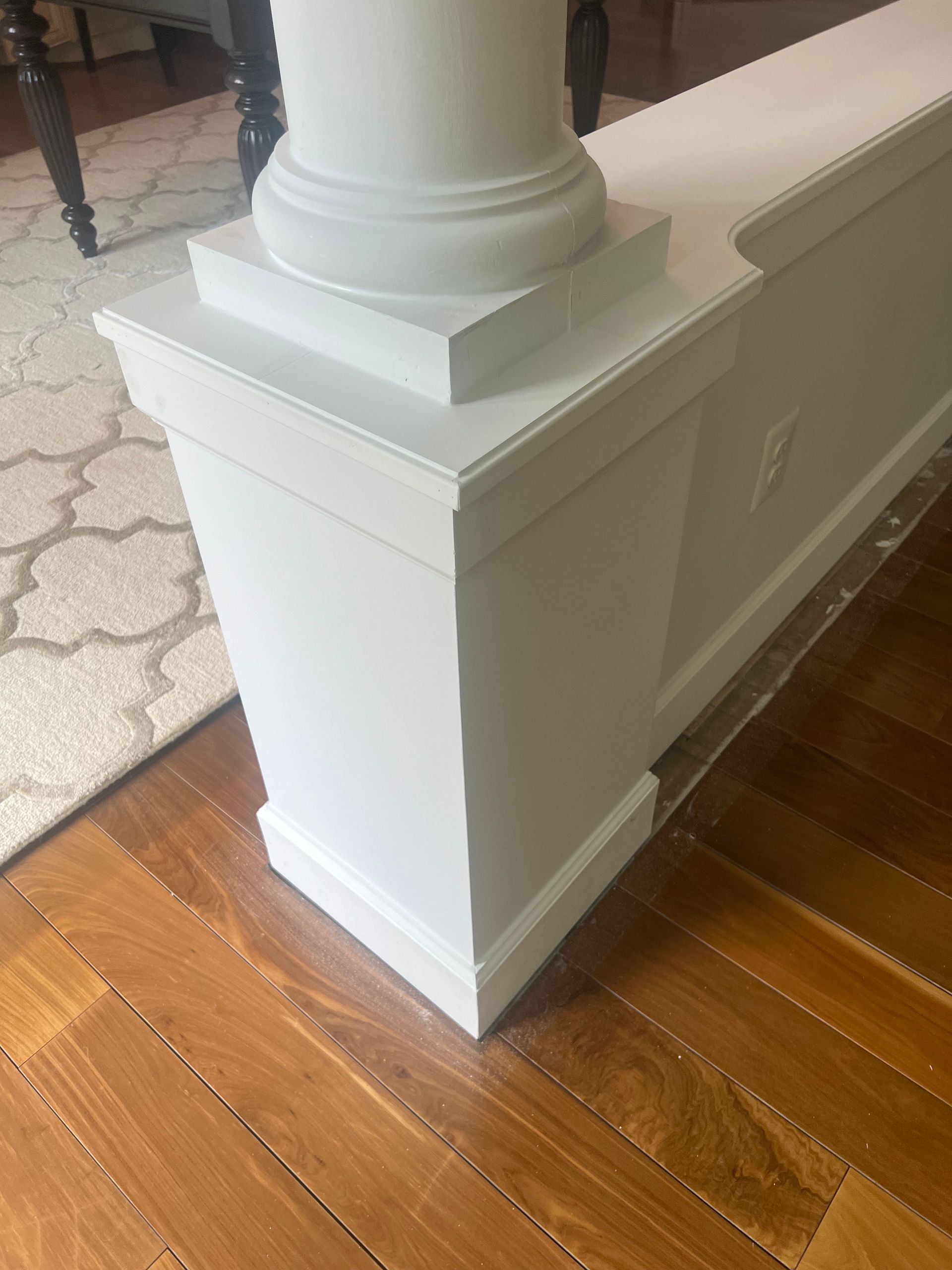 White wooden stair railing base beside a hardwood floor and patterned rug indoors