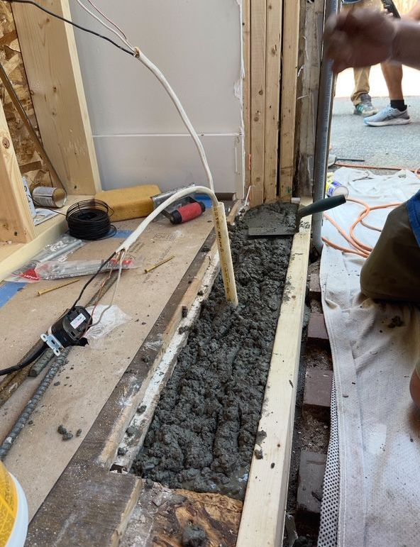 Construction site: cement poured into a wooden frame. Wires, tools, and workers in the background.