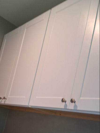 White kitchen cabinets with silver knobs installed over a light-colored wooden ledge against a gray wall.