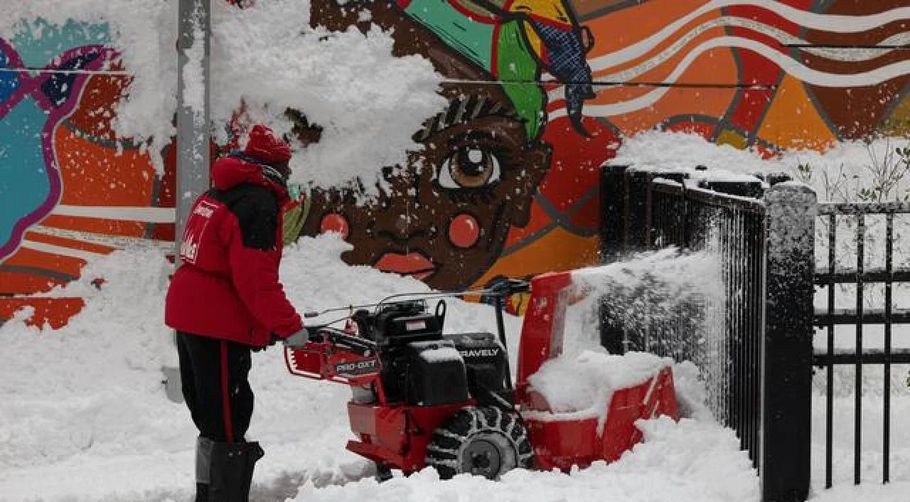 Person in red coat clearing snow beside a snowblower and red machine in front of a colorful mural and black fence