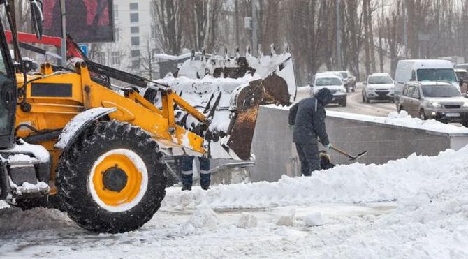 Yellow snowplow clearing a snowy street as a person shovels beside a wall in winter.