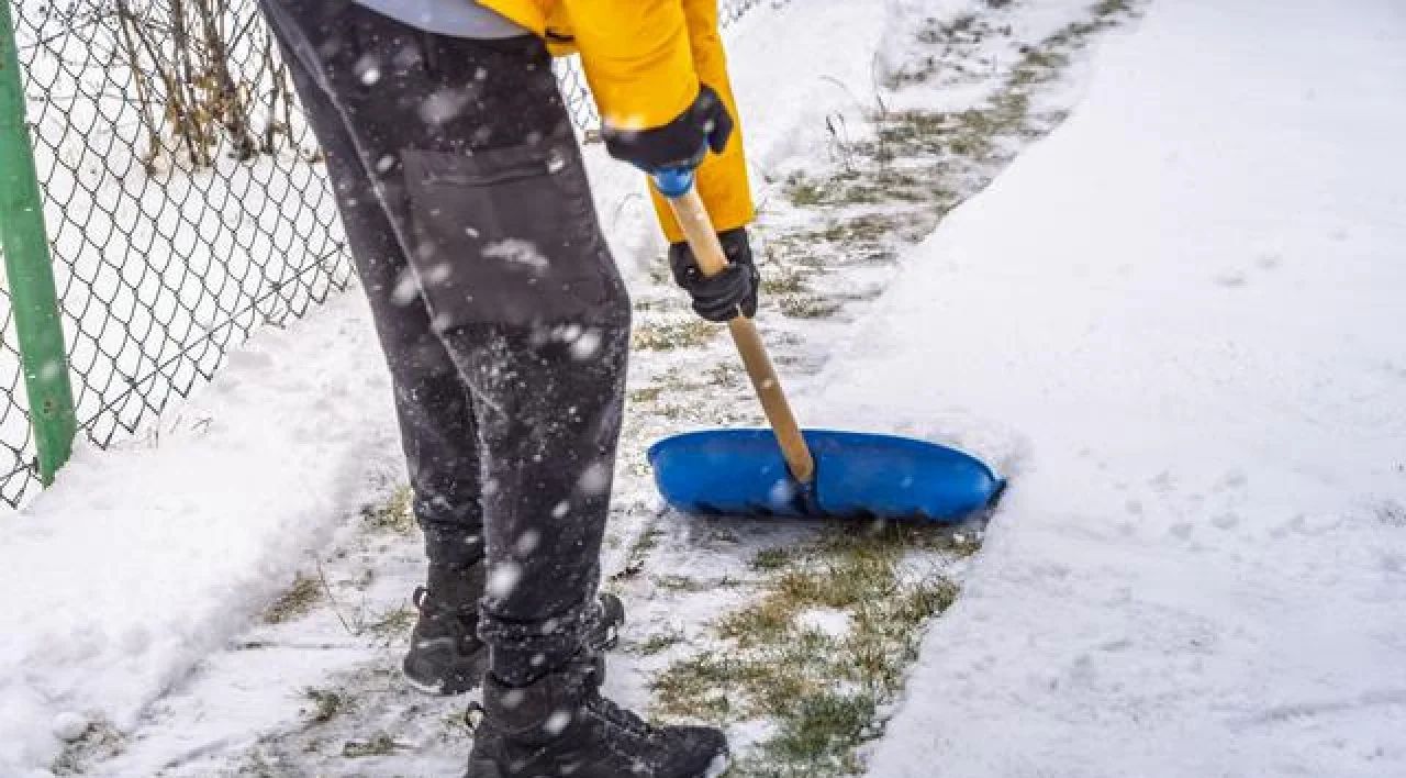 Person shoveling snow from a wet sidewalk near a chain-link fence