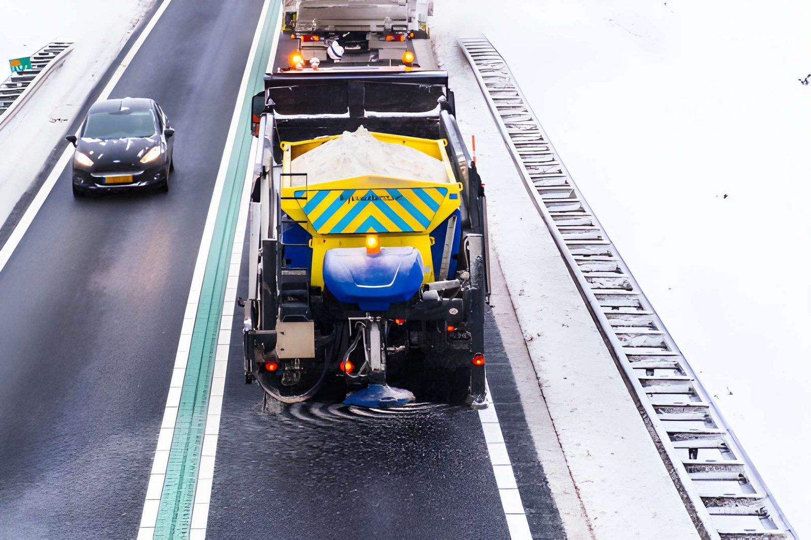 Road maintenance truck spraying liquid on a wet highway, with a car passing nearby.