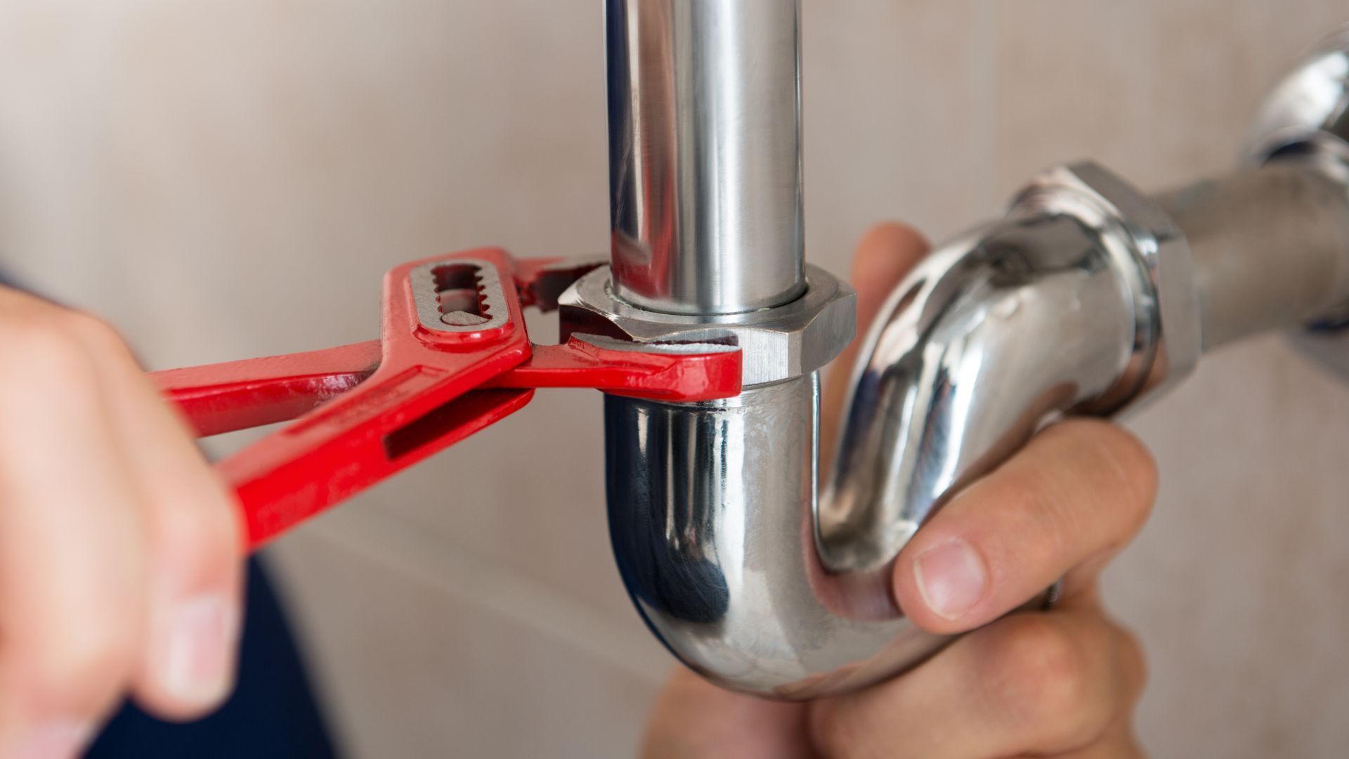 Person using a red wrench to tighten a chrome sink pipe under a bathroom sink