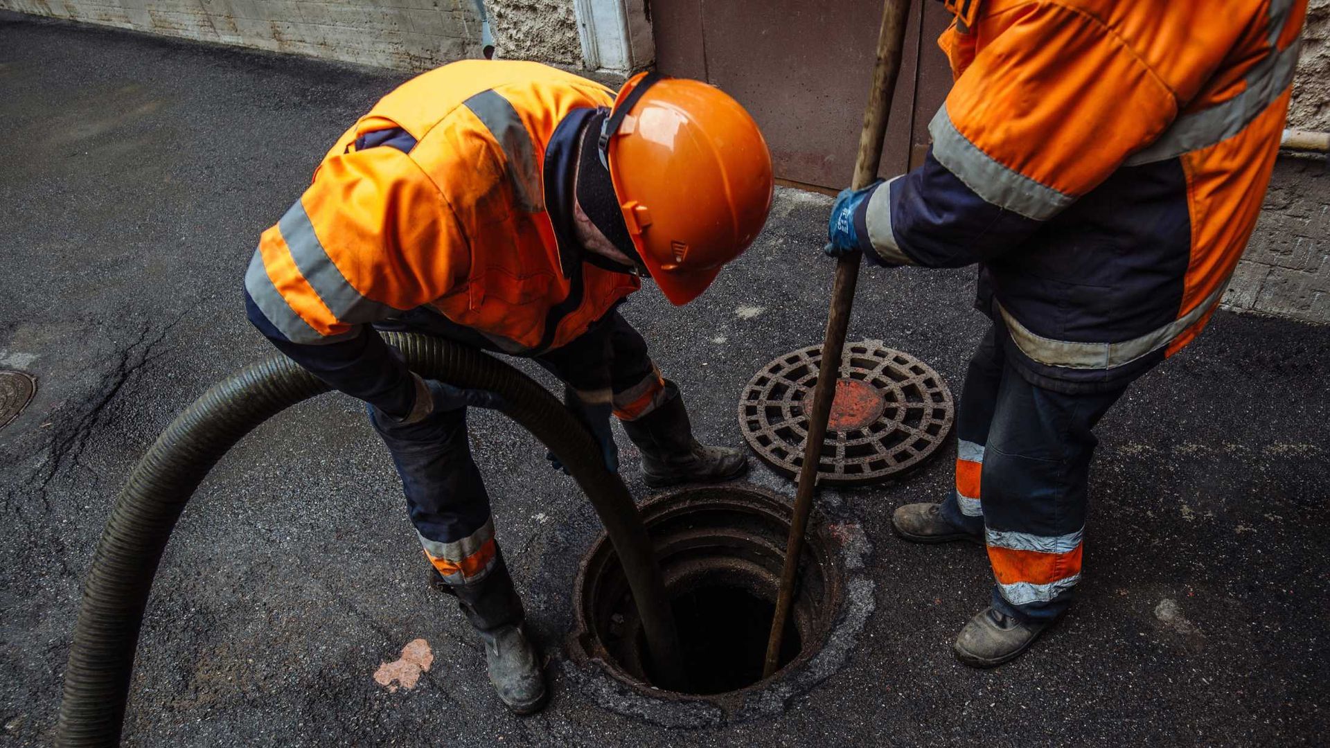 Two utility workers in orange safety gear inspecting a manhole on a wet street