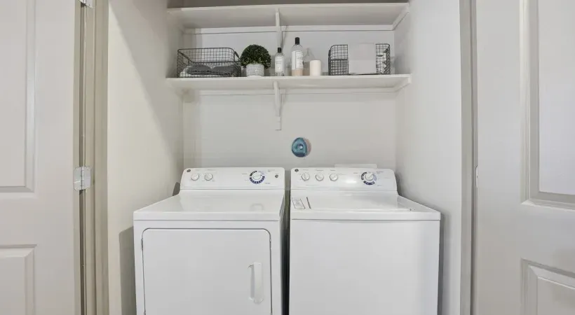 White laundry room with a washer and dryer side by side beneath a shelf at Layers Galleria.