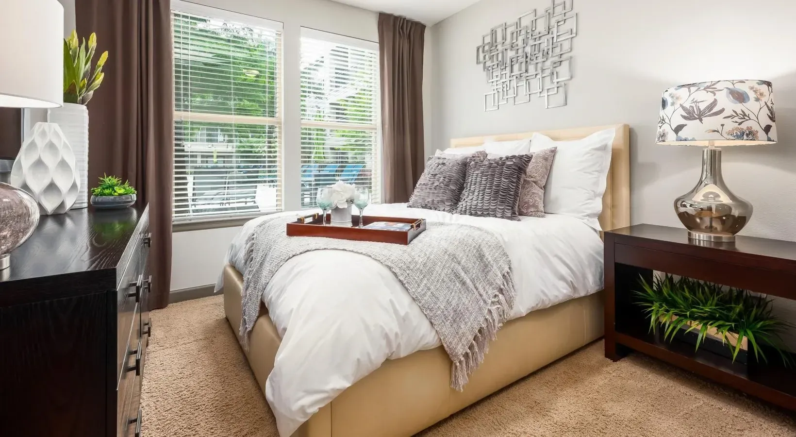 Cozy bedroom with beige bed, gray bedding, window blinds, and a bedside lamp next to a wooden dresser at Layers Galleria.