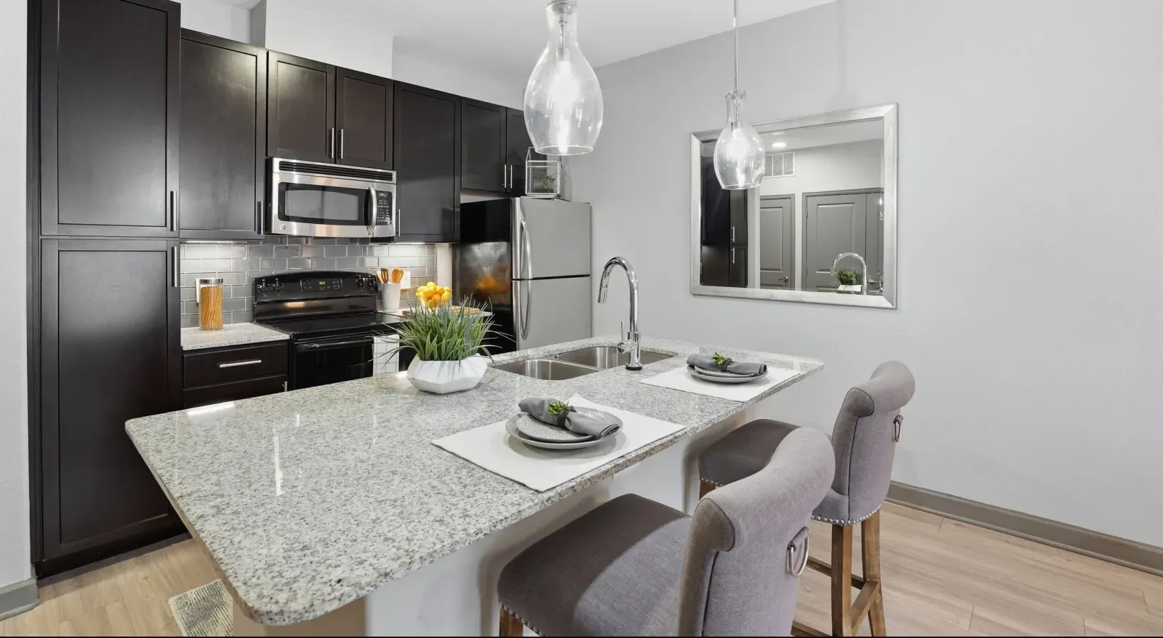 Modern kitchen with dark cabinets, granite island, pendant light, and two upholstered stools at Layers Galleria.