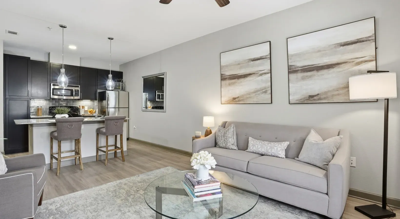 Modern open-concept living room with gray sofa, glass coffee table, floor lamp, and kitchen island in background at Layers Galleria.