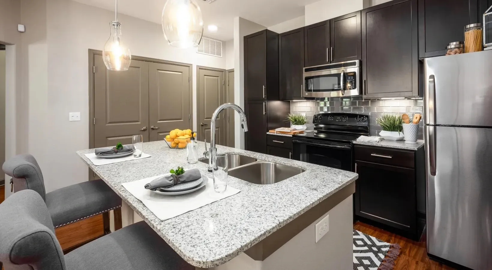Modern kitchen with dark cabinets, granite island, pendant lights, and gray upholstered chairs at Layers Galleria.