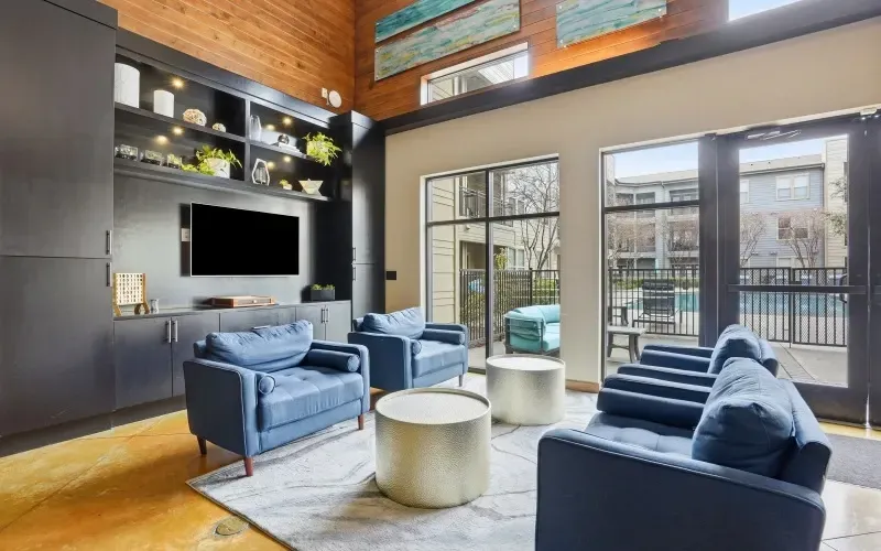 Modern living room with blue chairs, white coffee table, wall-mounted TV, and tall windows letting in natural light at Layers Galleria.