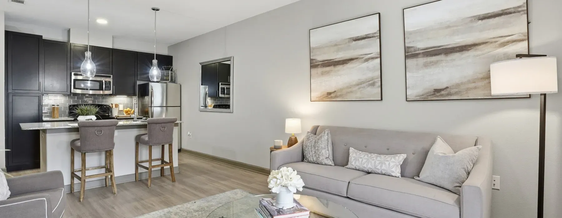 Bright open-plan living room with gray sofa, wall art, floor lamp, and kitchen bar stools in the background at Layers Galleria.