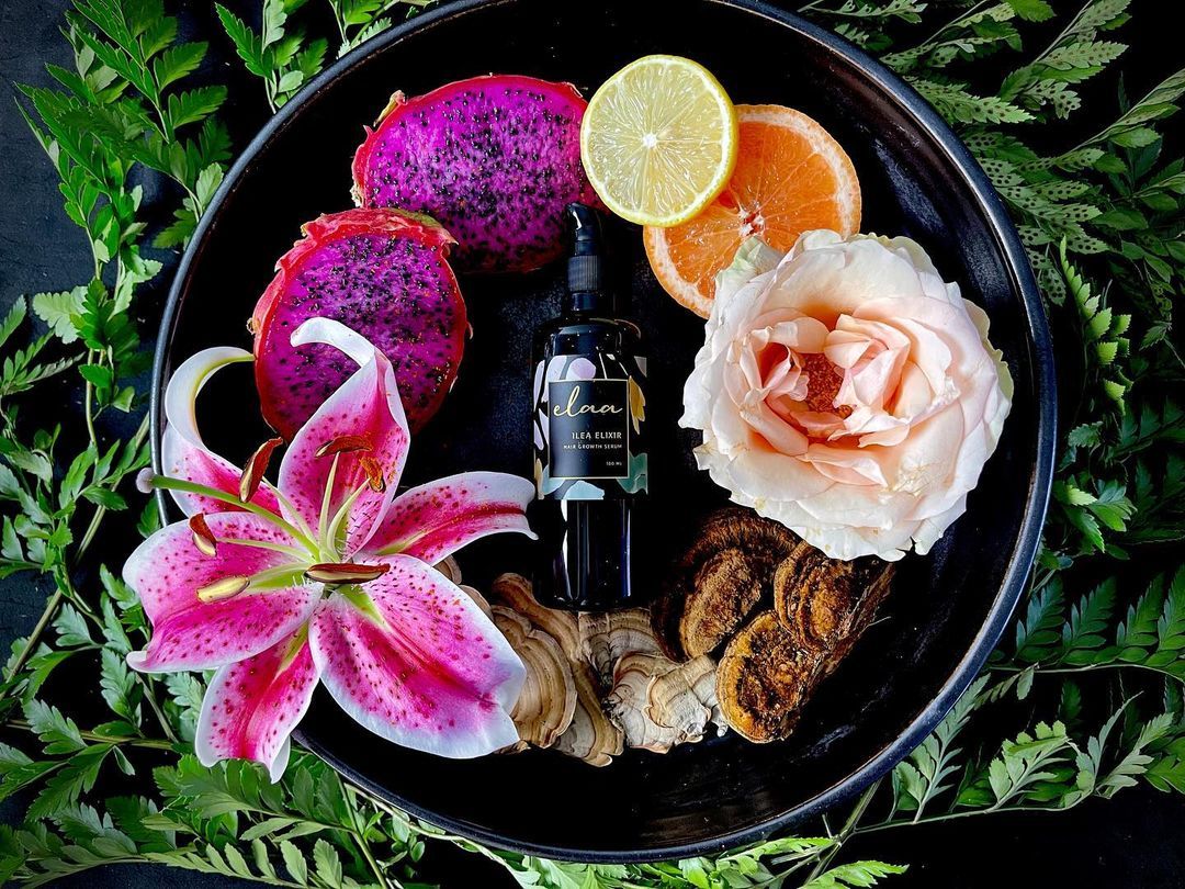 Black plate of tropical flowers, citrus slices, dragon fruit, and a small candle on greenery backdrop