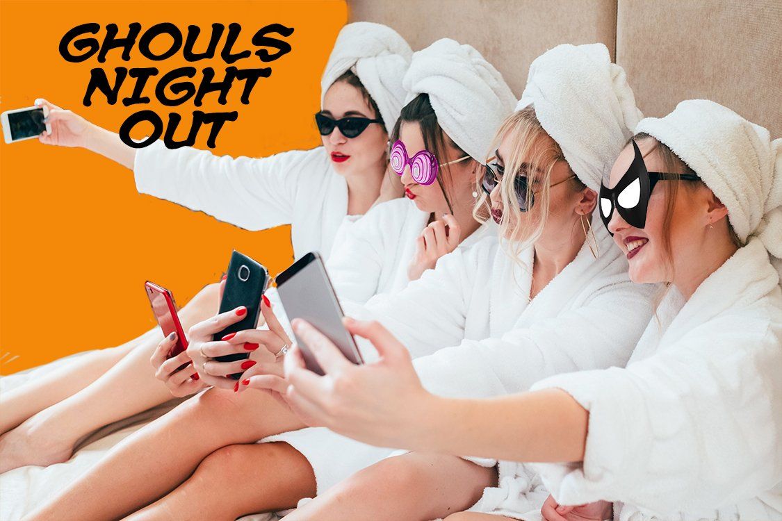 Four women in white robes and towel wraps taking a group selfie on a couch, wearing sunglasses.