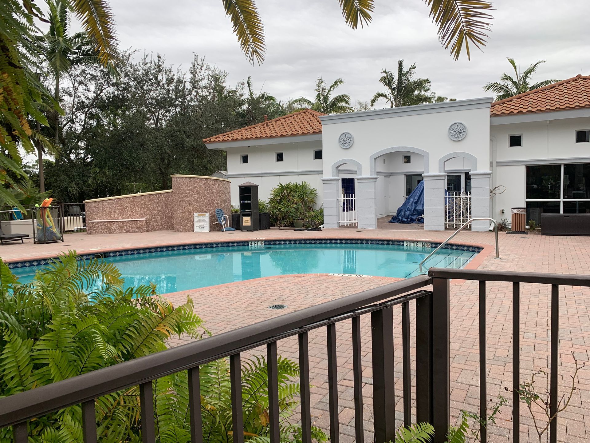 Resort-style pool with white building, terracotta roof, and patio seating behind a black railing.