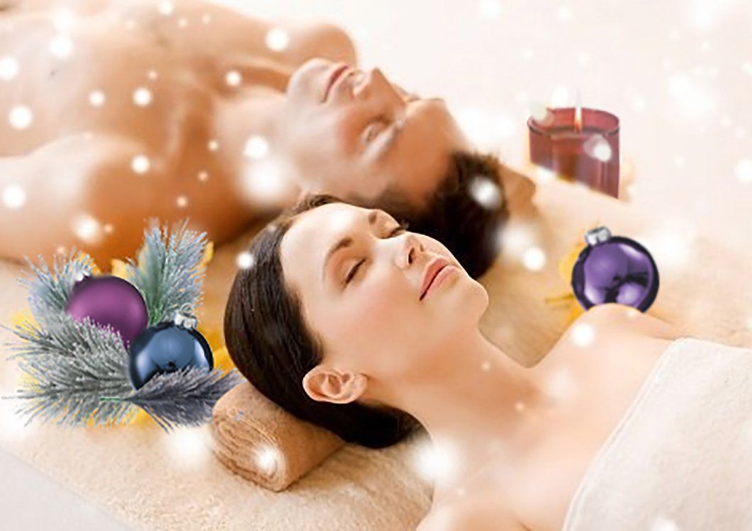 Two people relaxing in a warm spa with candles, bath salts, and holiday ornaments.