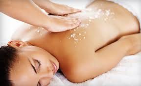 Person receiving a back massage with salt scrub on a white towel-covered spa bed