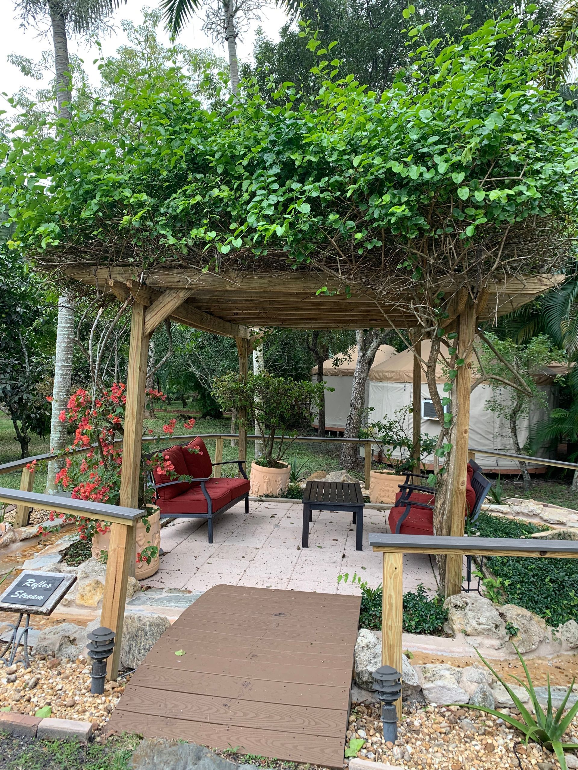 Garden pergola with leafy roof, red lounge chair, and small tables on a stone patio.