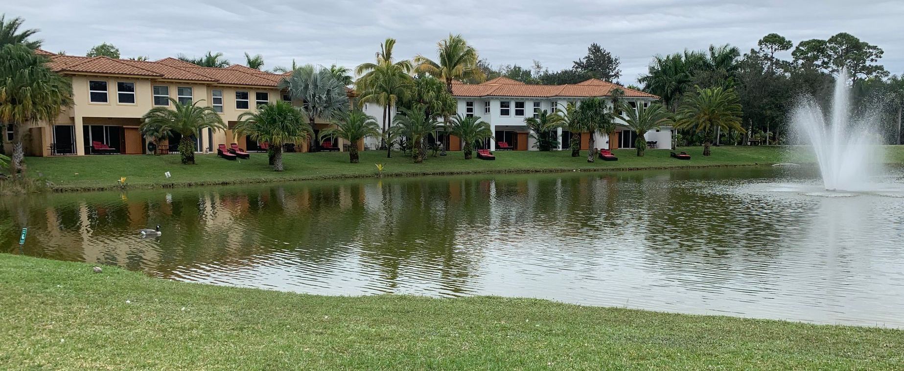 Lakeside homes with red tile roofs reflected in a pond, with a fountain spraying on the right.