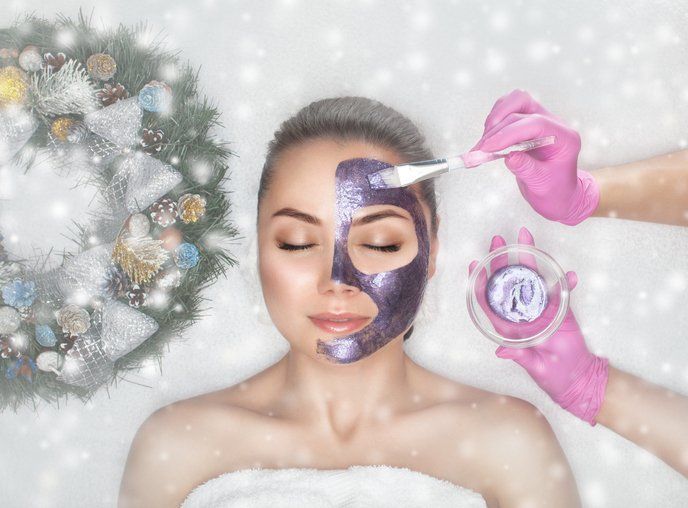 Spa treatment with woman receiving purple facial mask applied by gloved hands, with winter decor nearby