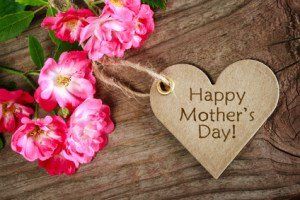 Pink flowers beside a wooden heart tag reading “Happy Mother’s Day” on rustic wood.