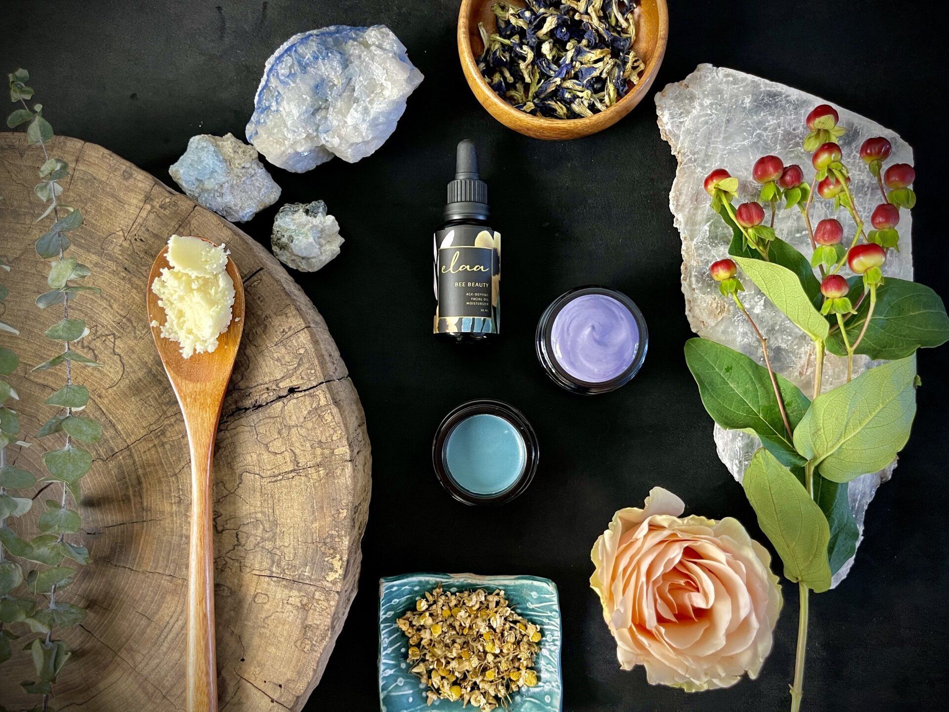 Natural skincare ingredients arranged on a rustic table with leaves, flowers, oils, and crystals
