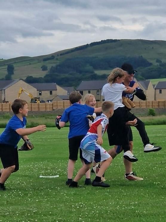 Children playing a game on a green field. One child is carried on another's back, others chase. Cloudy sky, hills.