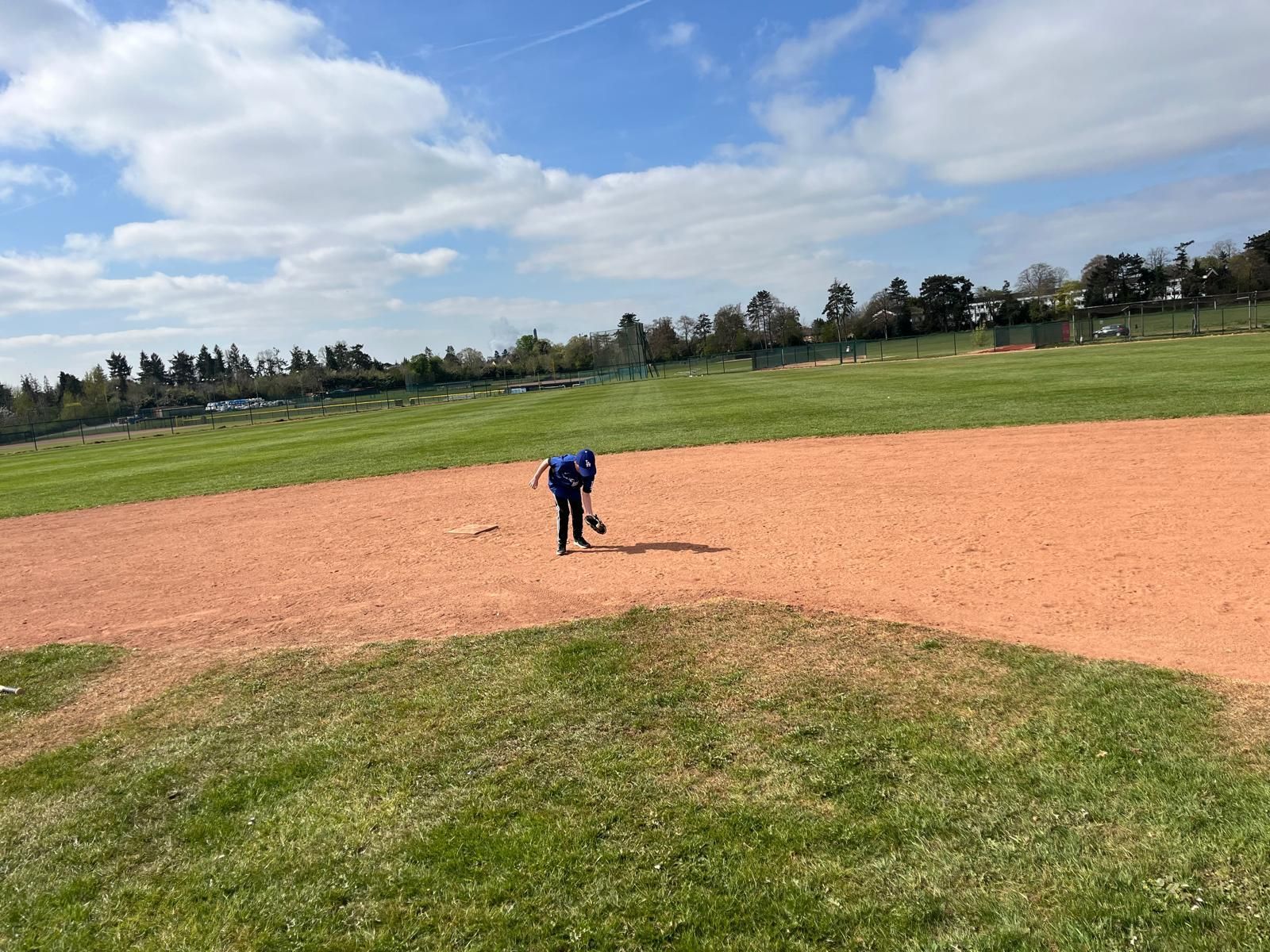 Person on baseball infield, raking dirt. Sunny day at a park, green grass, blue sky.