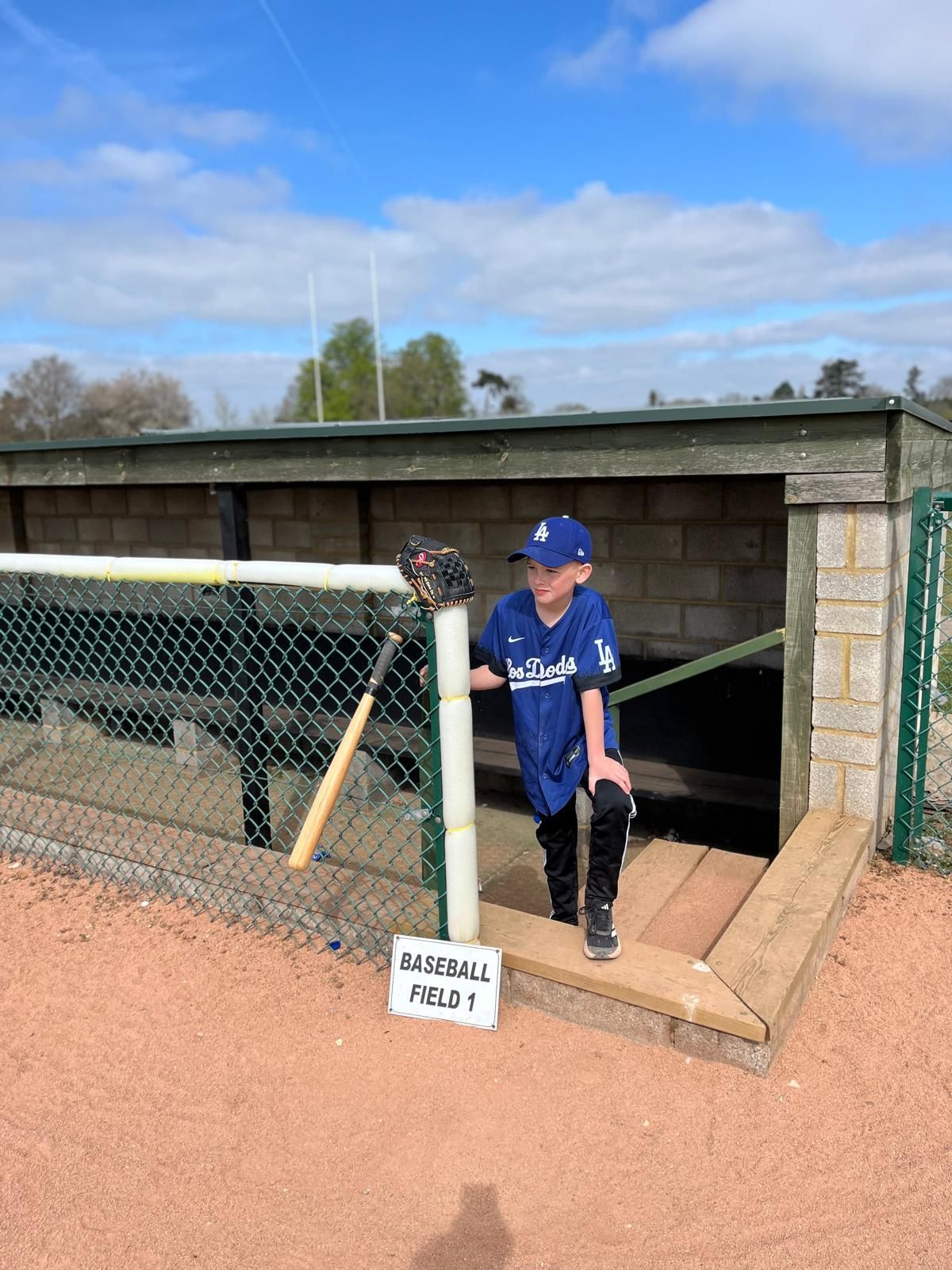 Child in Dodgers jersey stands in dugout with baseball glove and bat. Baseball field sign.