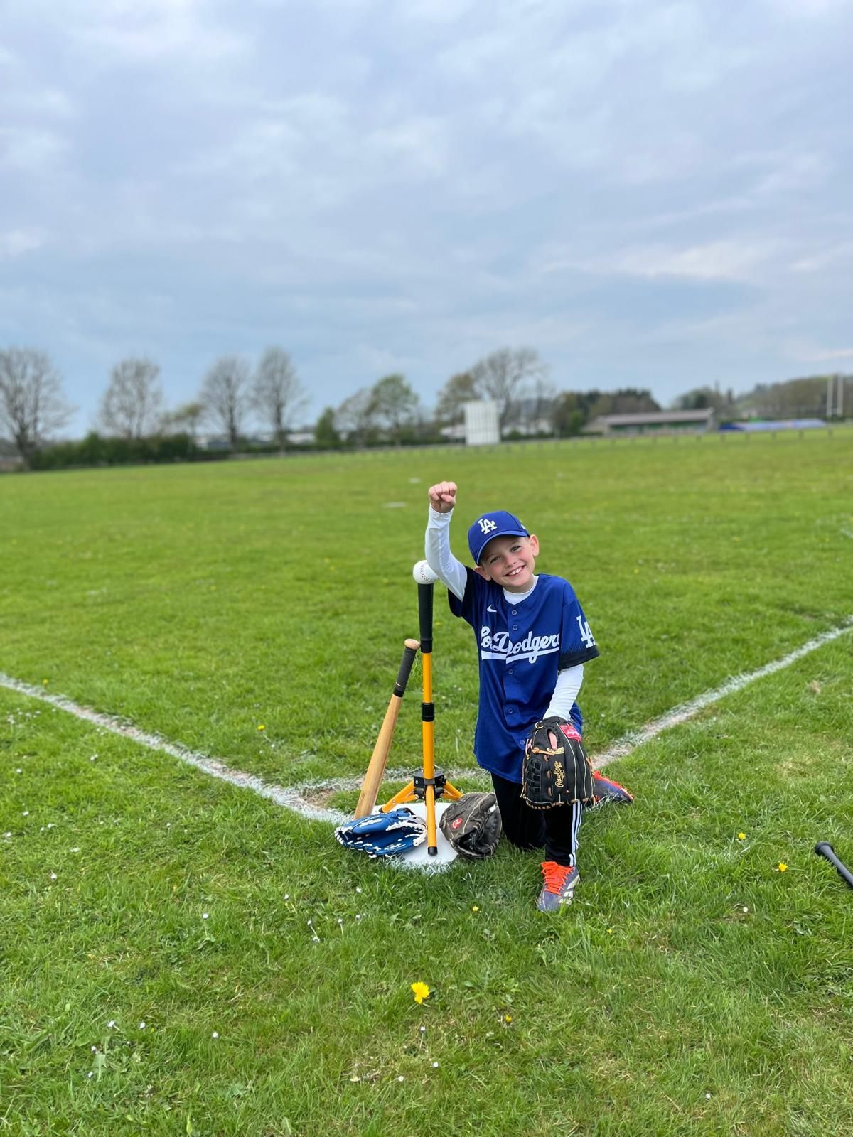 Young baseball player kneels on field, fist raised, near tee, bat, and glove. Blue jersey, green grass.