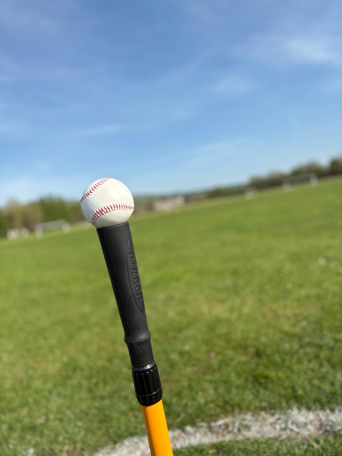 Baseball on a batting tee on a grassy field, blue sky in the background.