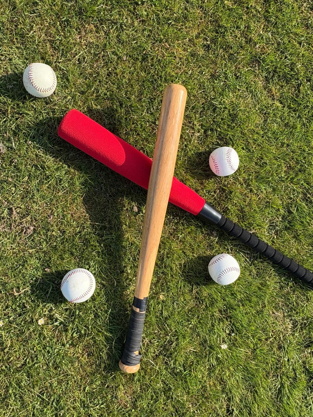 Two baseball bats and four baseballs on grass. One bat is red, one is wooden.