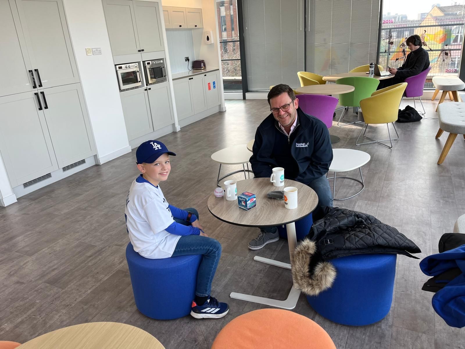 A man and Arthur sit at a table in a modern room, smiling. Arthur wears a baseball cap and jersey.