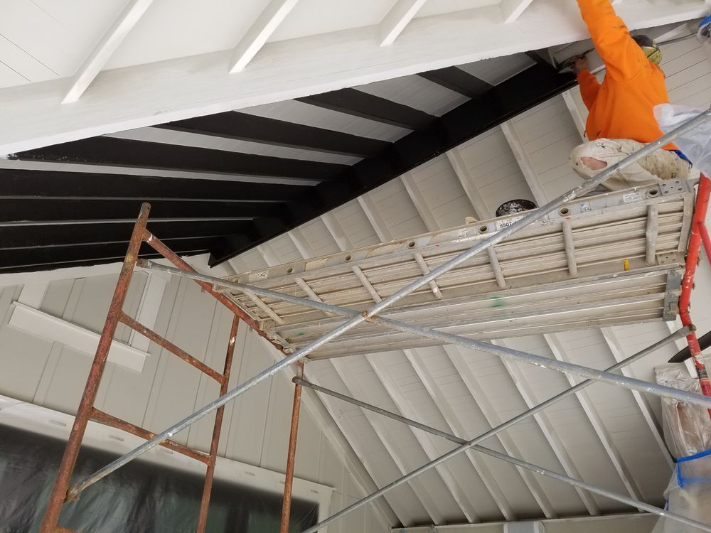 Person in orange painting a black ceiling, standing on a scaffolding. White trim around the ceiling.
