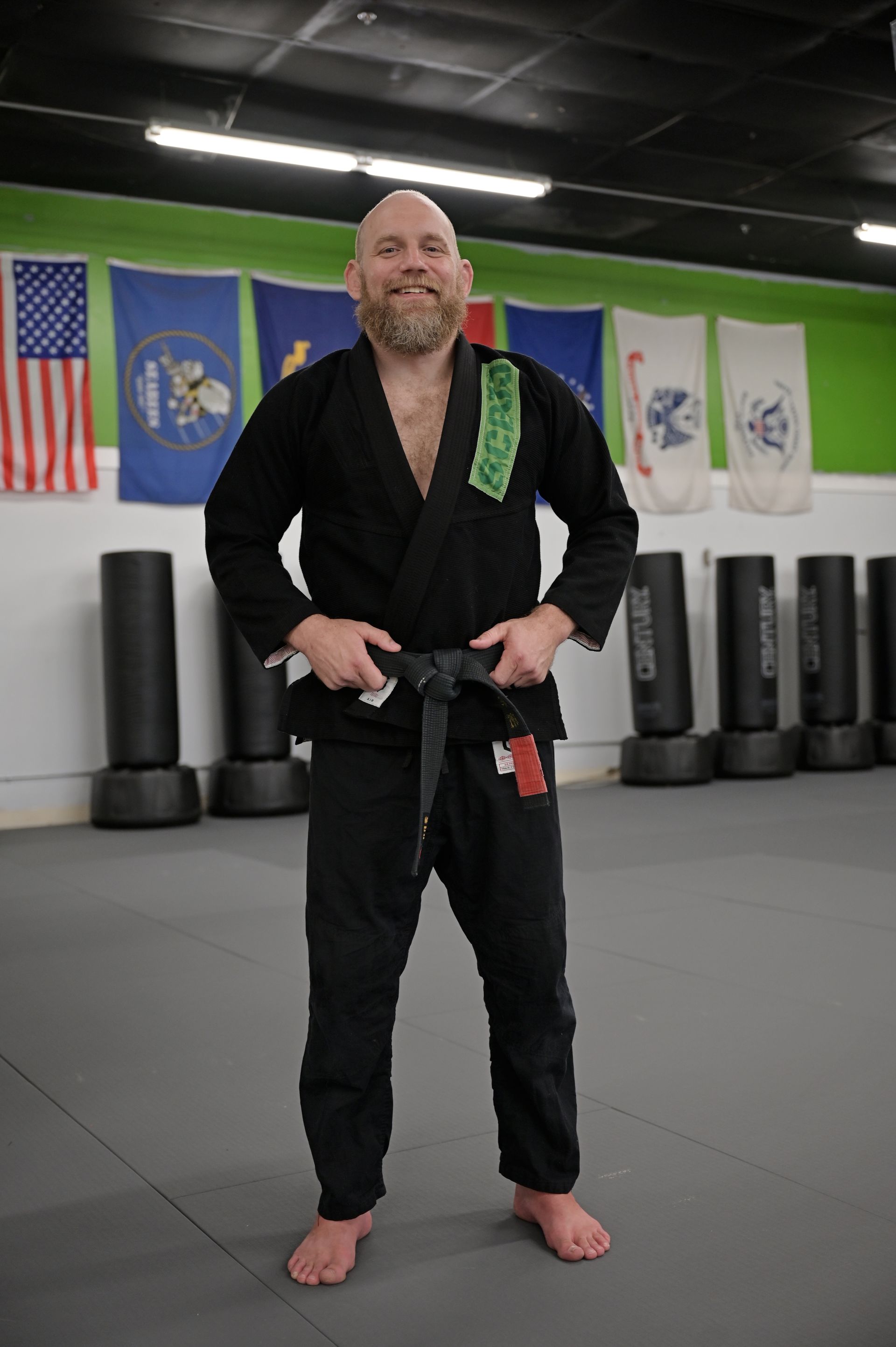 Martial arts instructor in black gi standing barefoot in a dojo with flags and punching bags behind him