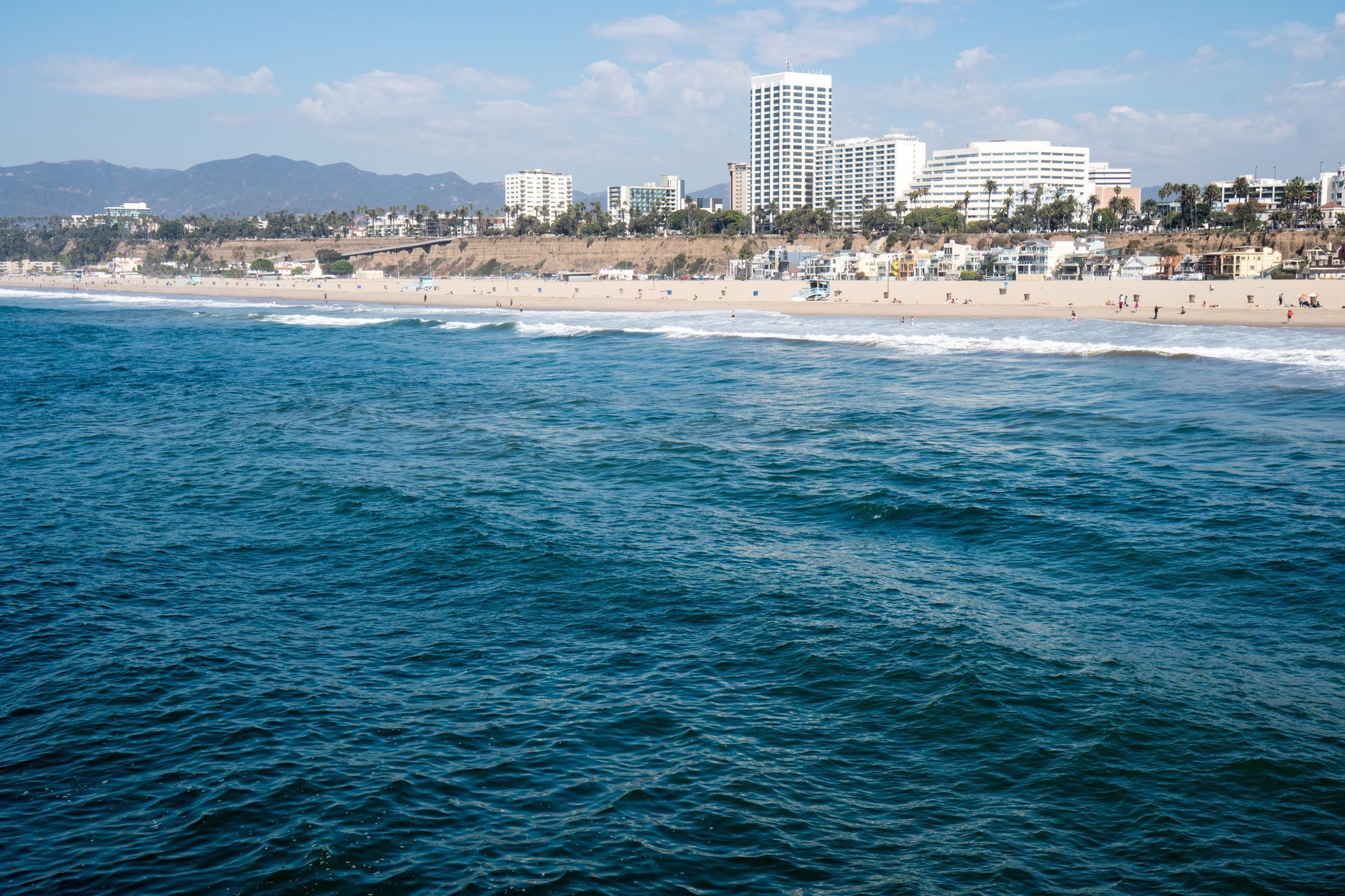 Blue ocean waves with a sandy beach and distant coastal buildings under a clear sky.