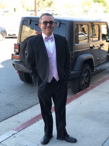 Man in black suit and pink tie standing beside a dark SUV on a city sidewalk