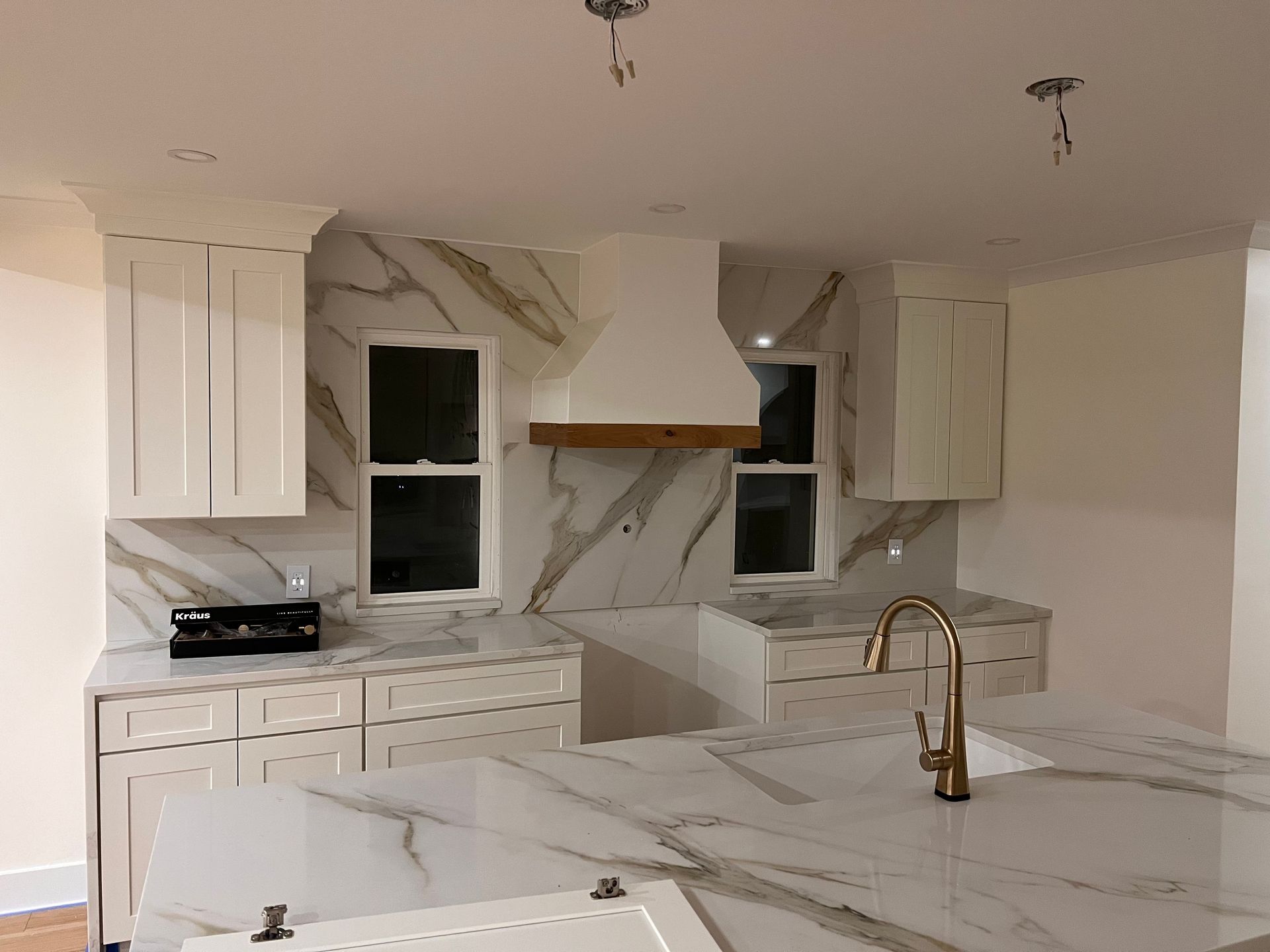White kitchen with marble backsplash, cabinets, and island. Gold faucet, wooden hood.