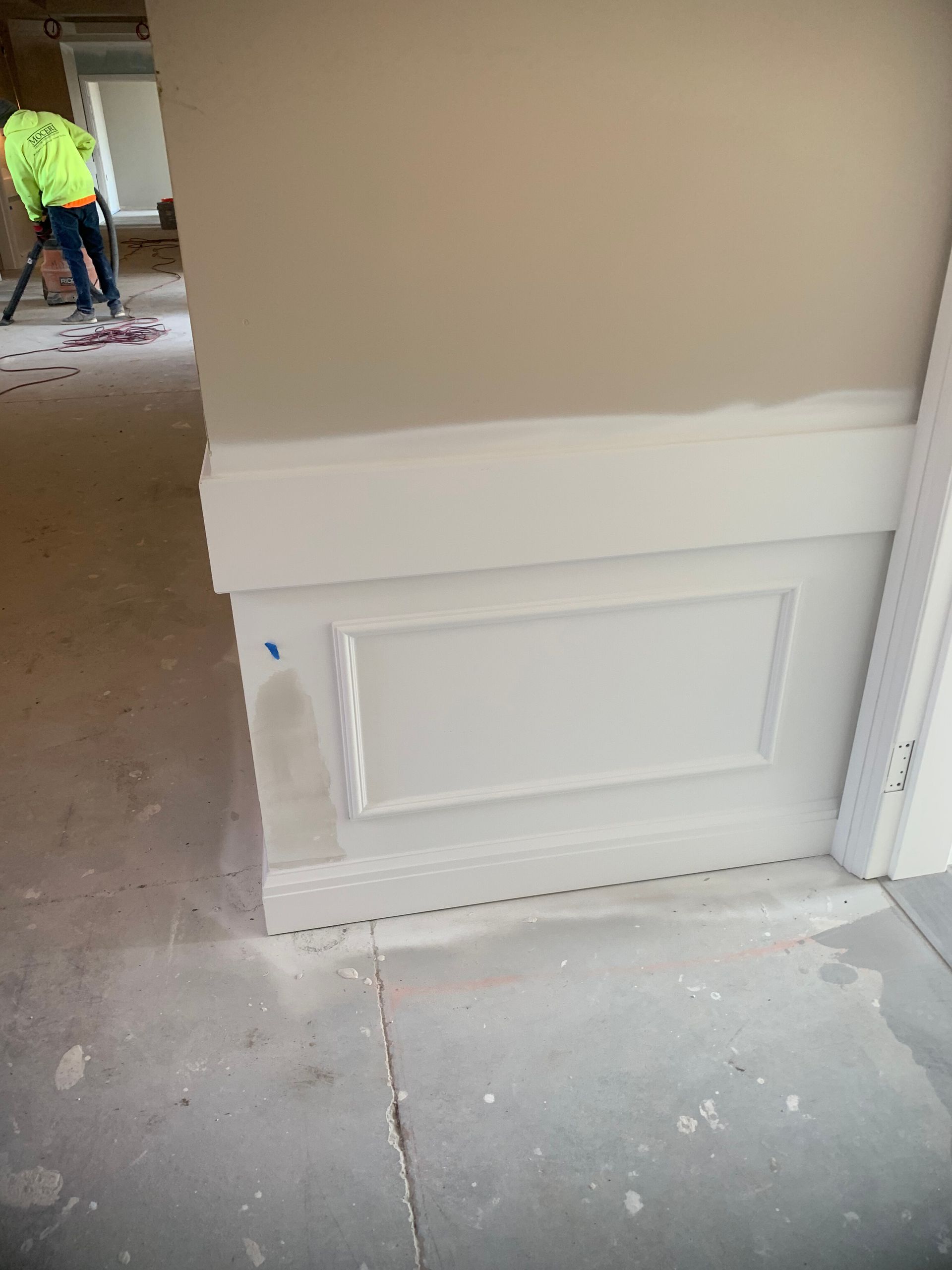 White wainscoting on beige wall in room under construction; person vacuuming in background.