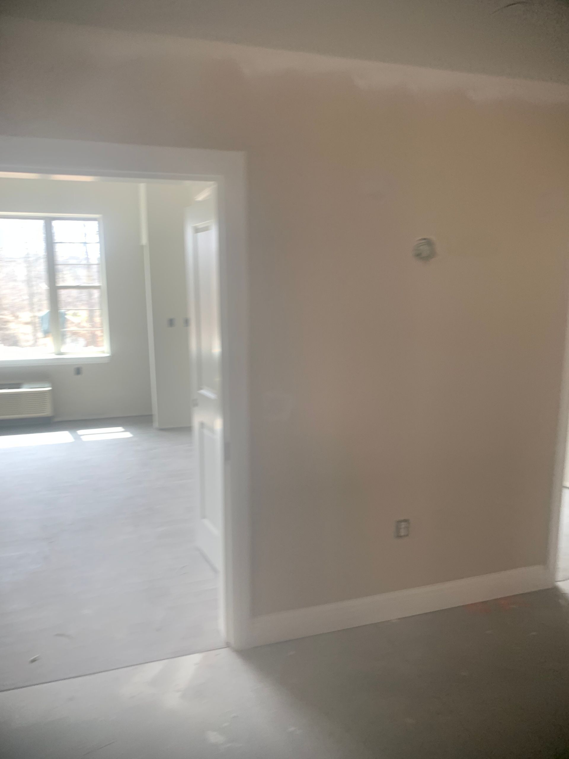 Interior view of a room with an open doorway, white trim, and a window in the background. Light beige walls.