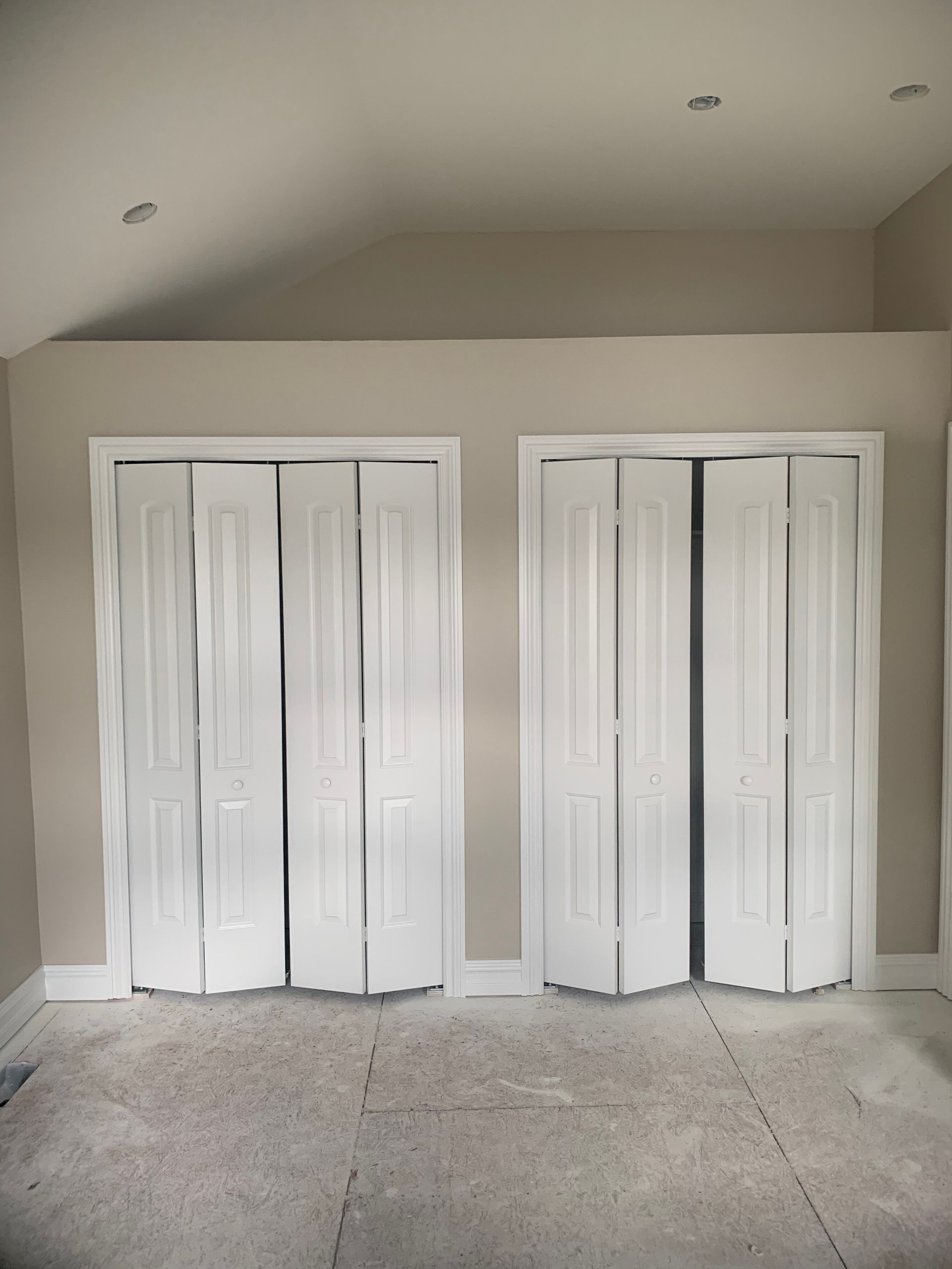 Two white folding closet doors in a room with light tan walls, cream trim, and a gray floor.