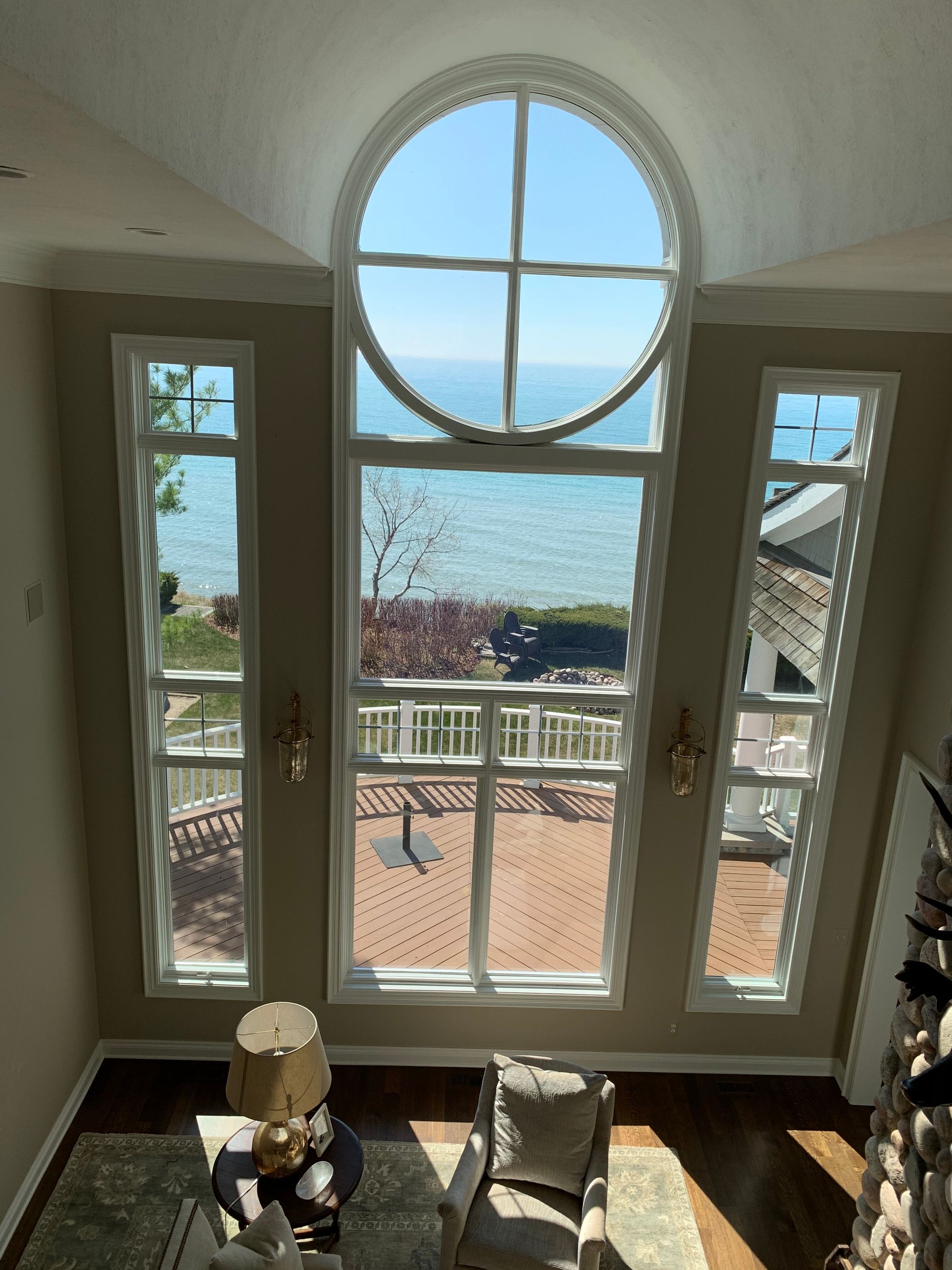 View of a lake through large windows. Tall arched window above rectangular windows, wooden deck below. Blue sky and water visible.