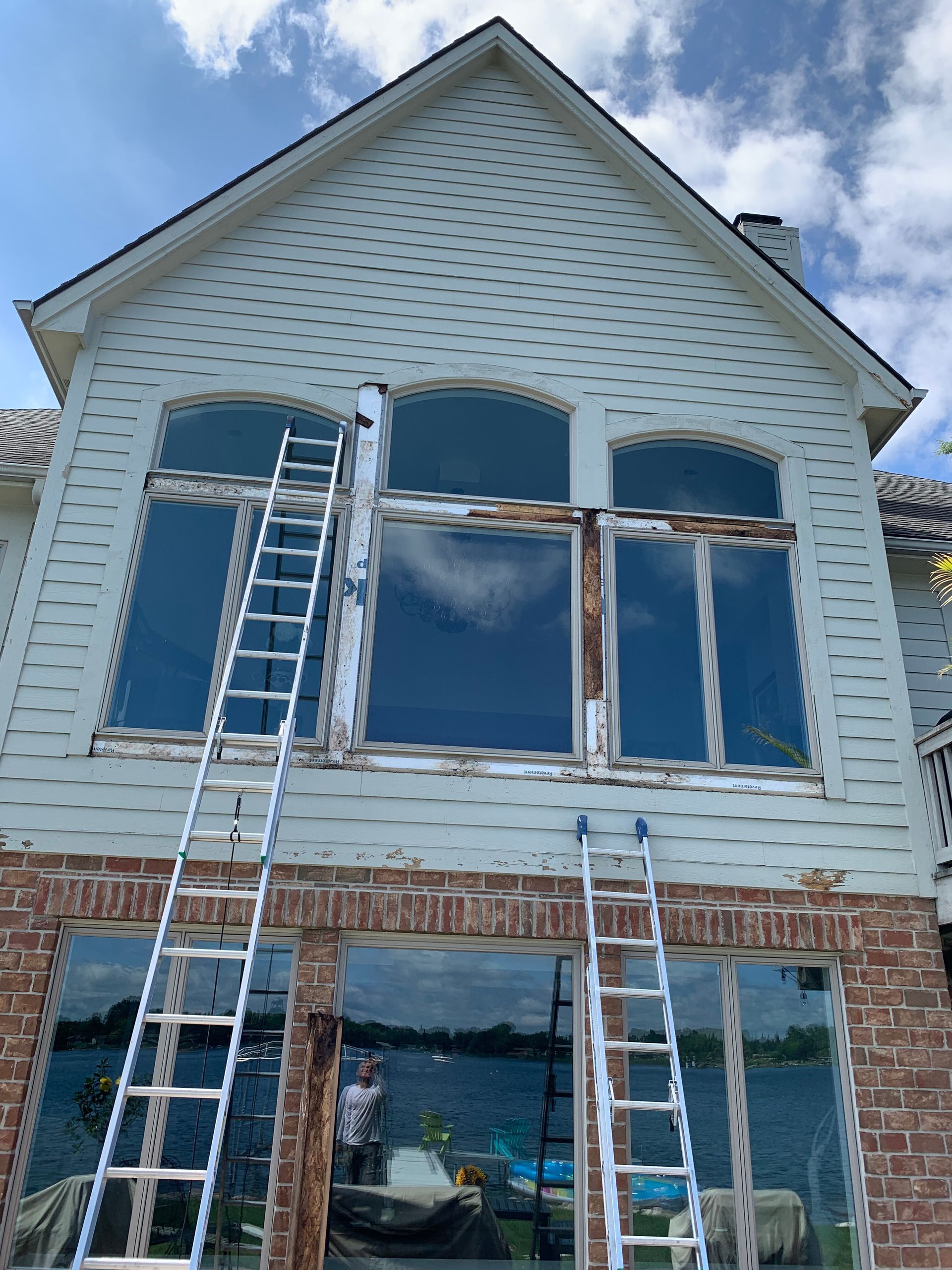 Exterior of a house with two ladders propped against the window frames. Siding is painted and peeling. Brickwork at the bottom.