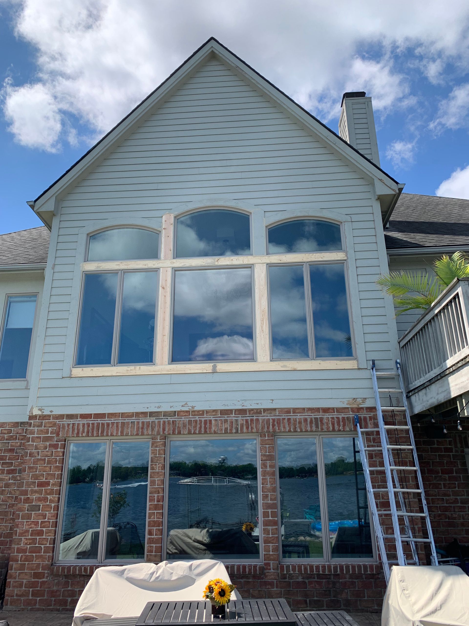 Two-story house with large windows, blue siding, and brick, reflected sky. Ladder leans against the house.