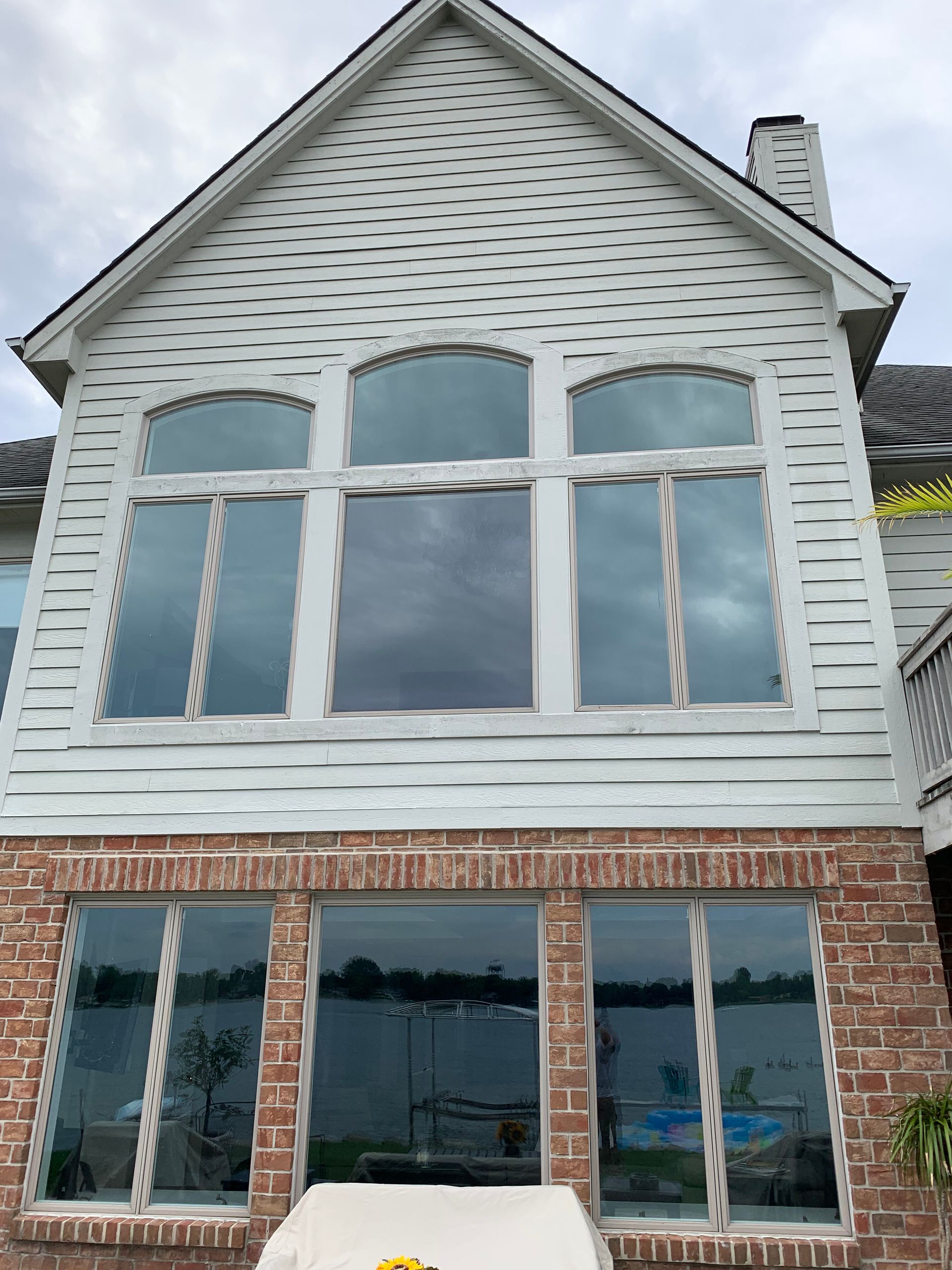 Two-story house with large windows reflecting cloudy sky. Red brick base, white siding, lake view.