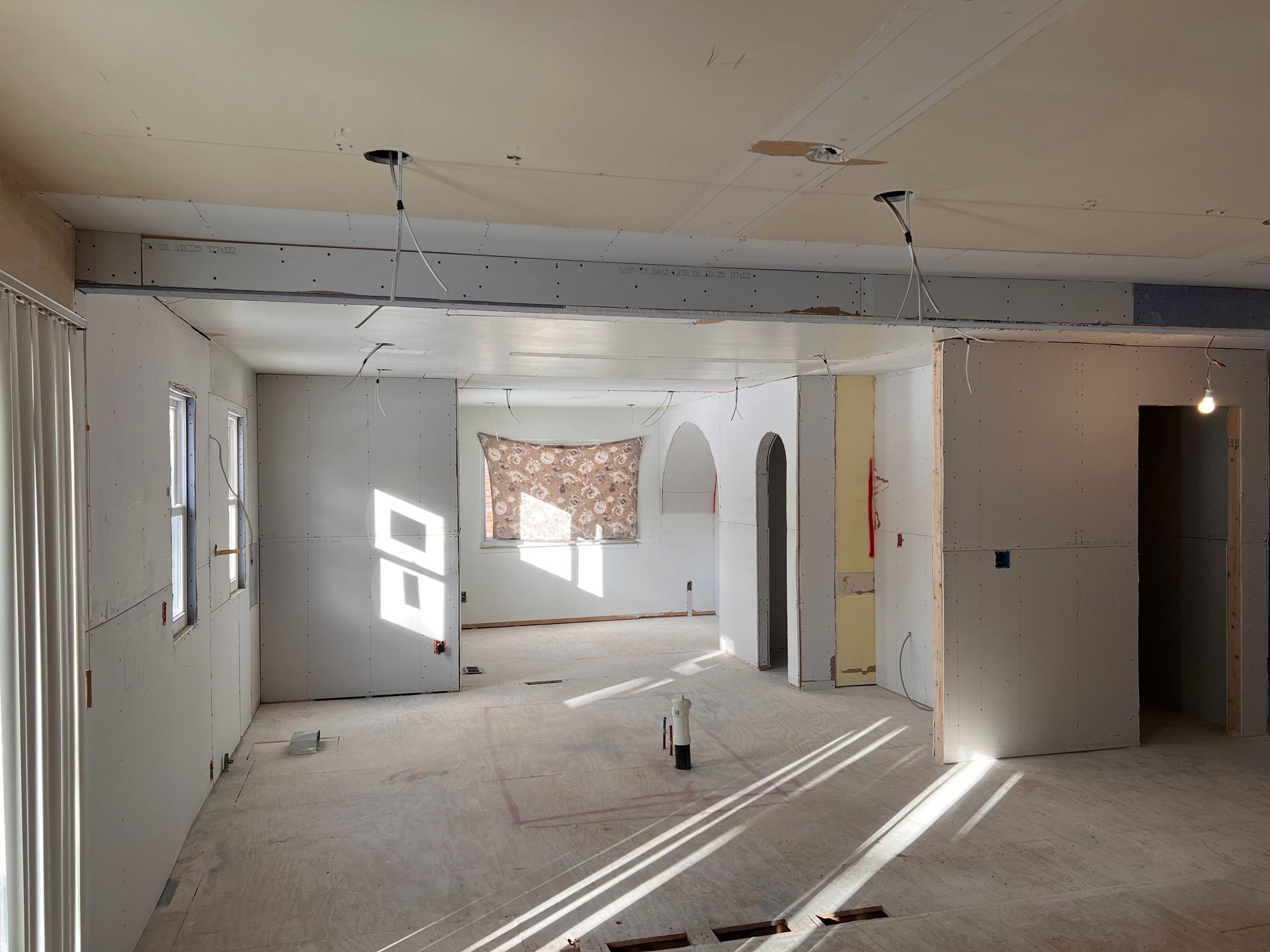 Interior of a room under construction, with exposed drywall, wiring, and architectural features. Sunlight streams through windows.