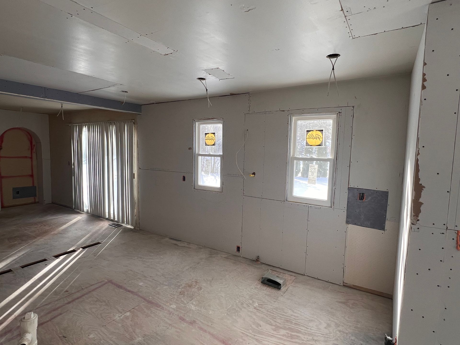 Interior of a room under construction with drywall on walls and ceiling, two windows, and a sliding glass door.