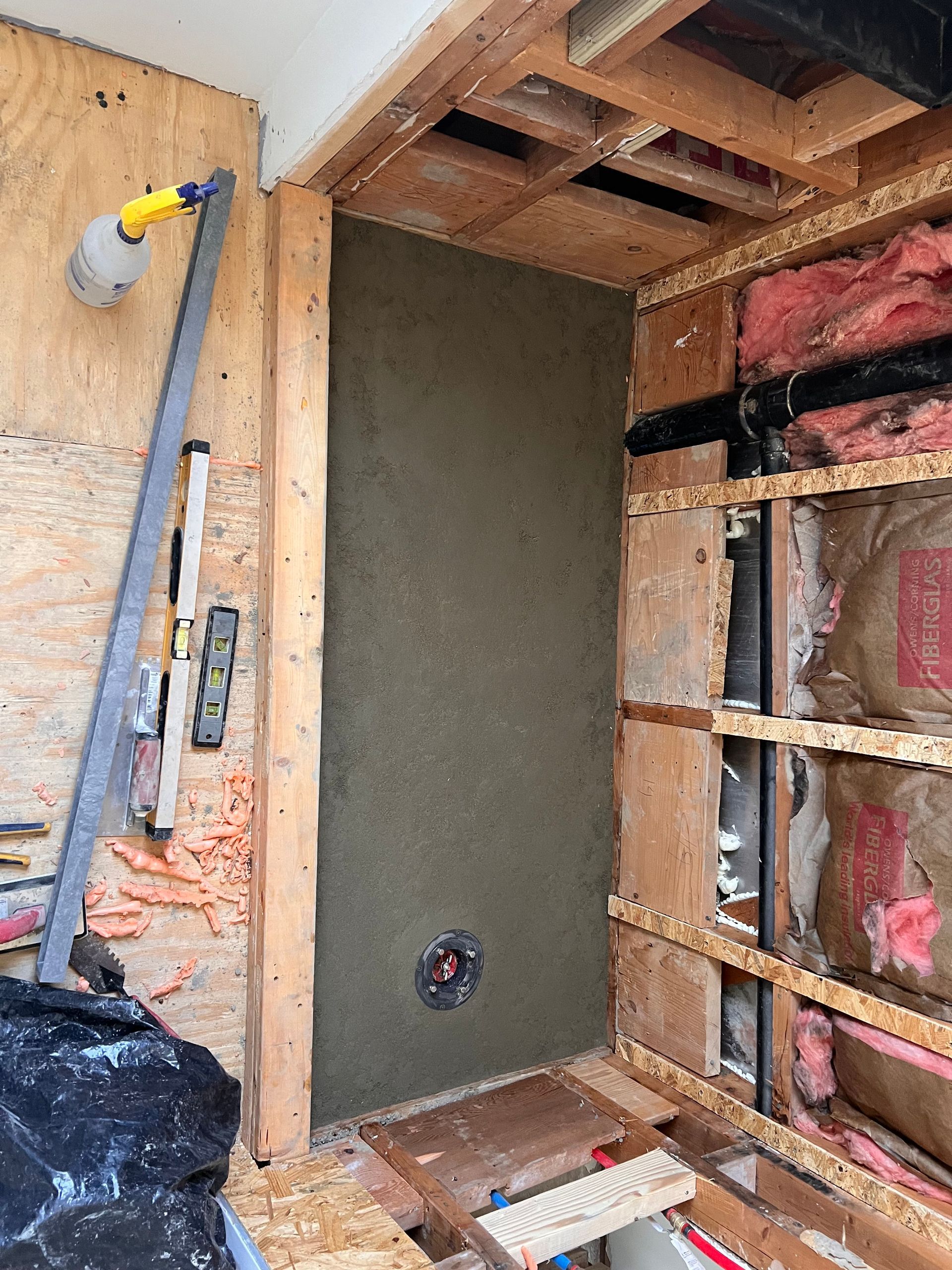 Shower wall construction in progress: cement-covered back wall, exposed wooden framing, tools, and pipes.