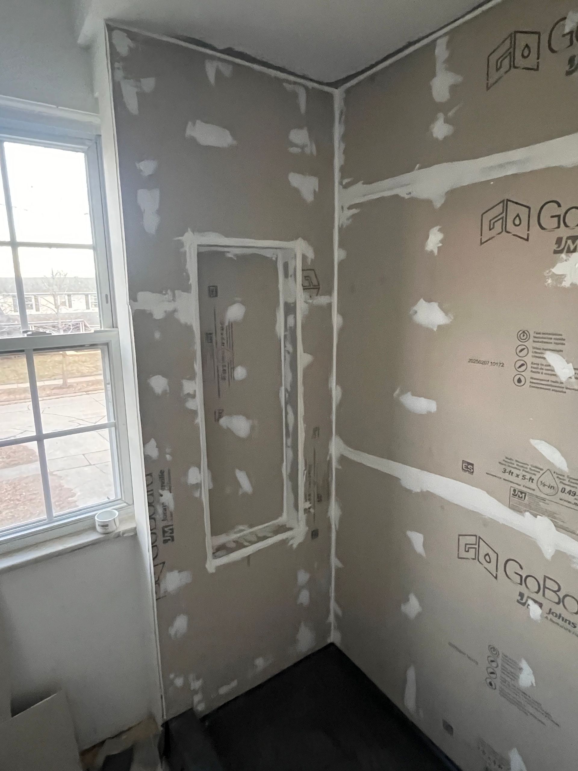 Bathroom walls under construction with shower niche. Beige wallboard and window visible.
